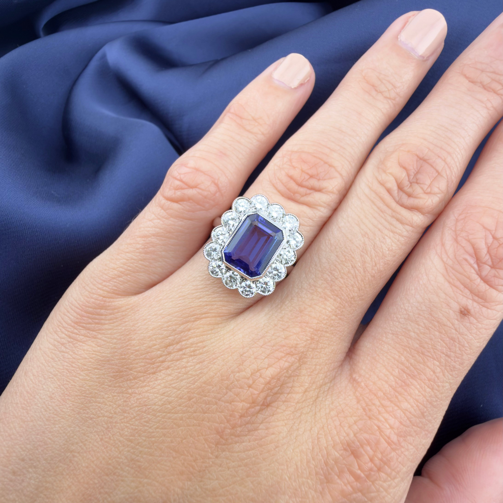 A close-up of a hand wearing a white gold ring with a rectangular deep purple Tanzanite surrounded by a halo of diamonds, set against a draped navy blue fabric background.